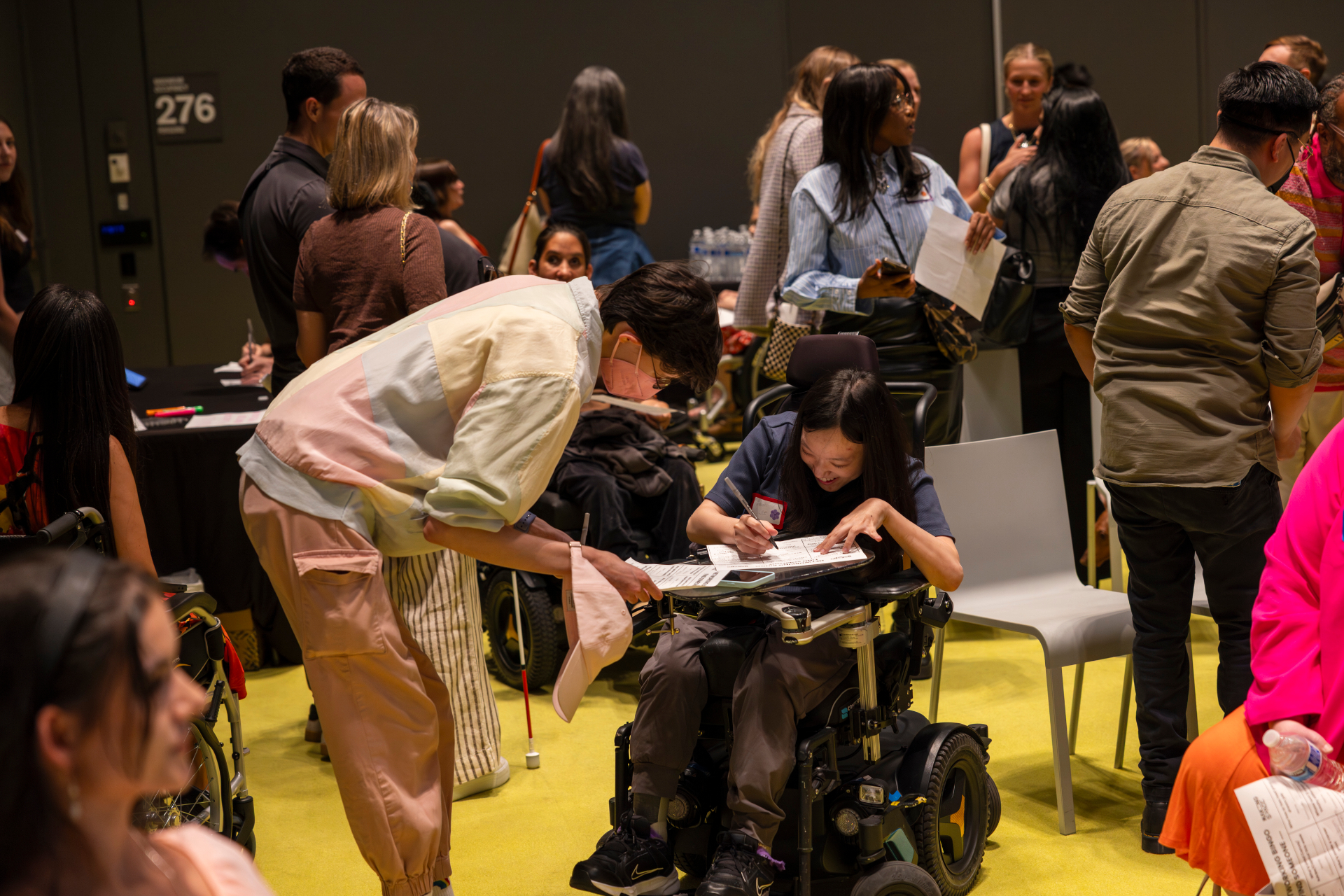 Two Making Space members in wheelchairs collaborate on a worksheet together amid a busy community event.