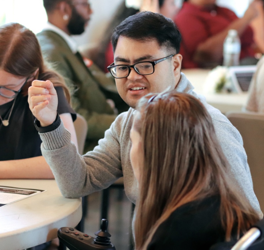 Marcus Reyes, a wheelchair user wearing glasses and a gray sweater, in conversation at a table.